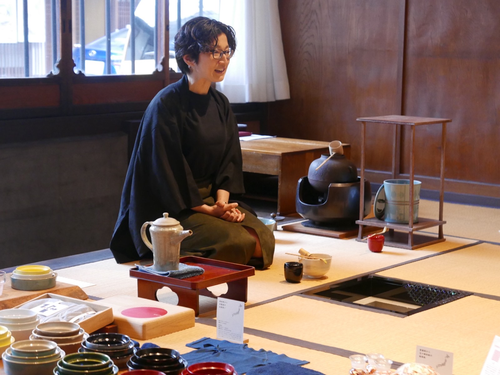 A host in dark robes sits on tatami beside an iron kettle, with tea bowls and crafts laid out in the foreground.