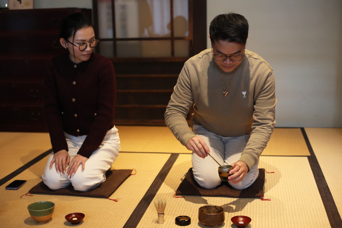 Two guests in white T-shirts sit on tatami, each whisking matcha in a glass bowl with a bamboo whisk.