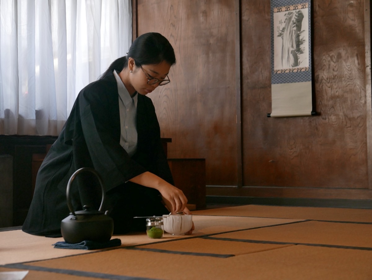 A host kneels on tatami beside an iron kettle, scooping matcha into a glass tea bowl.