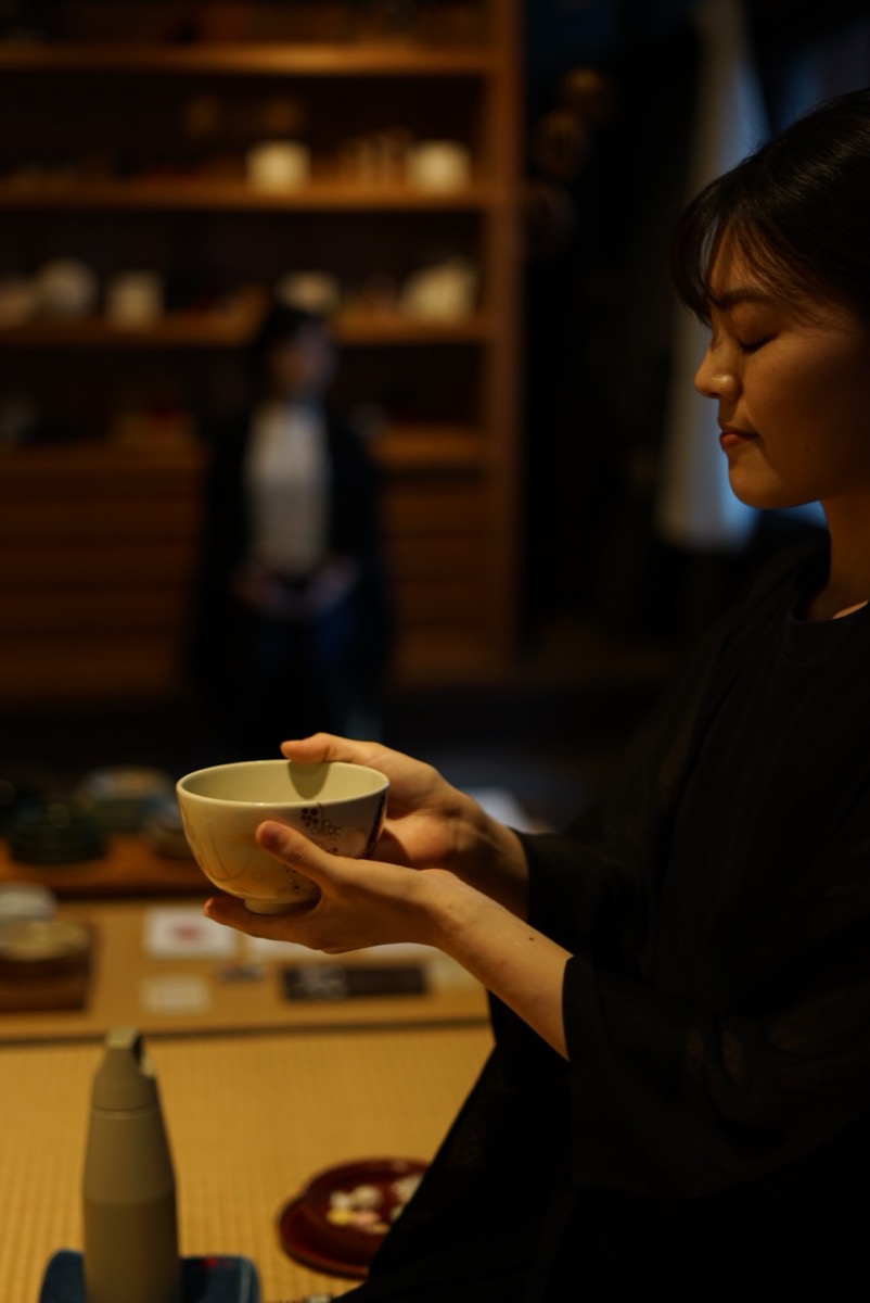 A host raises a glass tea bowl with both hands to drink, in front of shelves of crafts at aeru gojo.