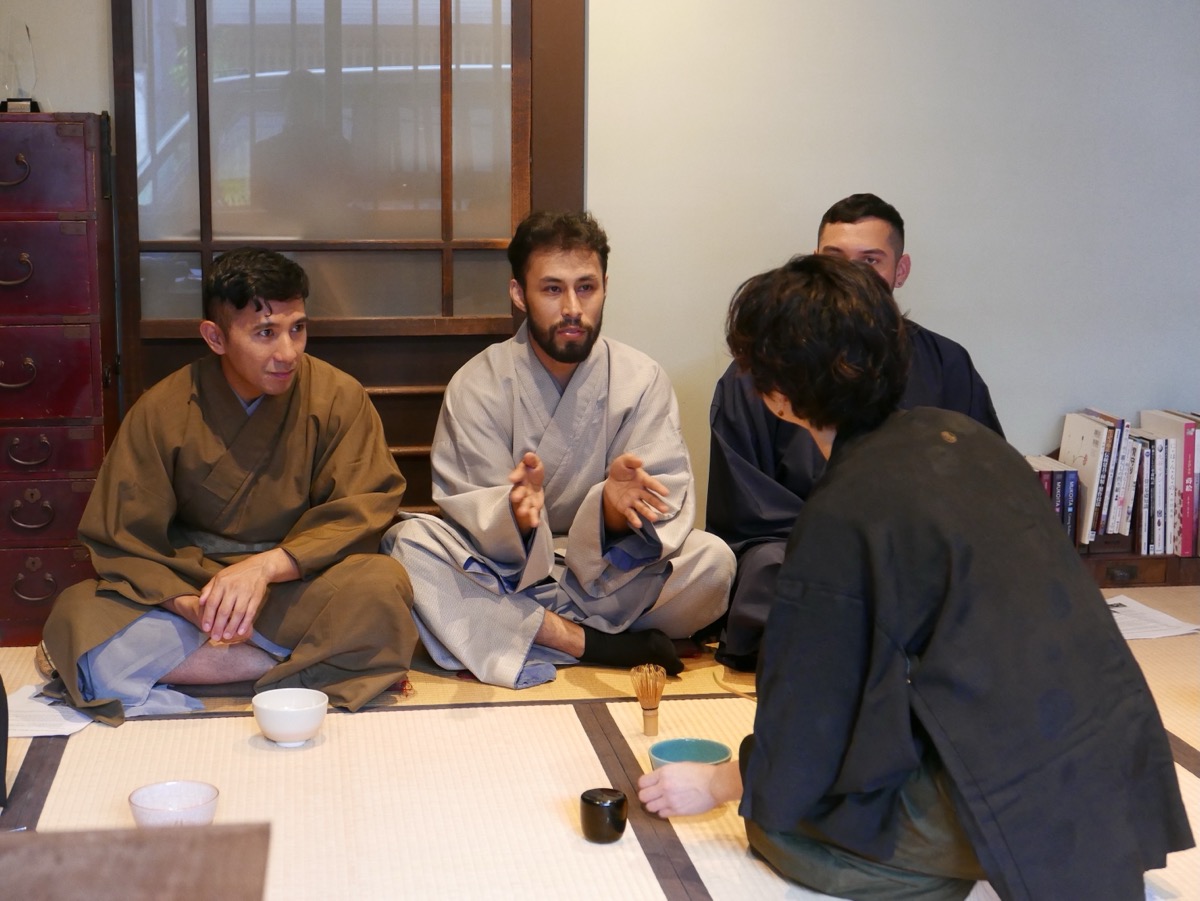 A host speaks with a gesture while four guests in summer clothing sit on tatami, listening.