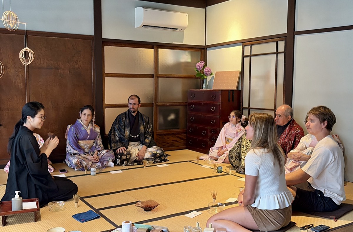 A host speaks to a group of guests seated on tatami, some in kimono and some in everyday summer clothes.