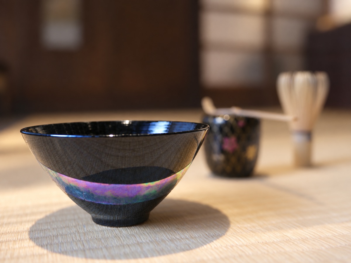 A white-and-gold glass tea bowl on tatami, with a glass caddy of matcha and a bamboo whisk beside it.