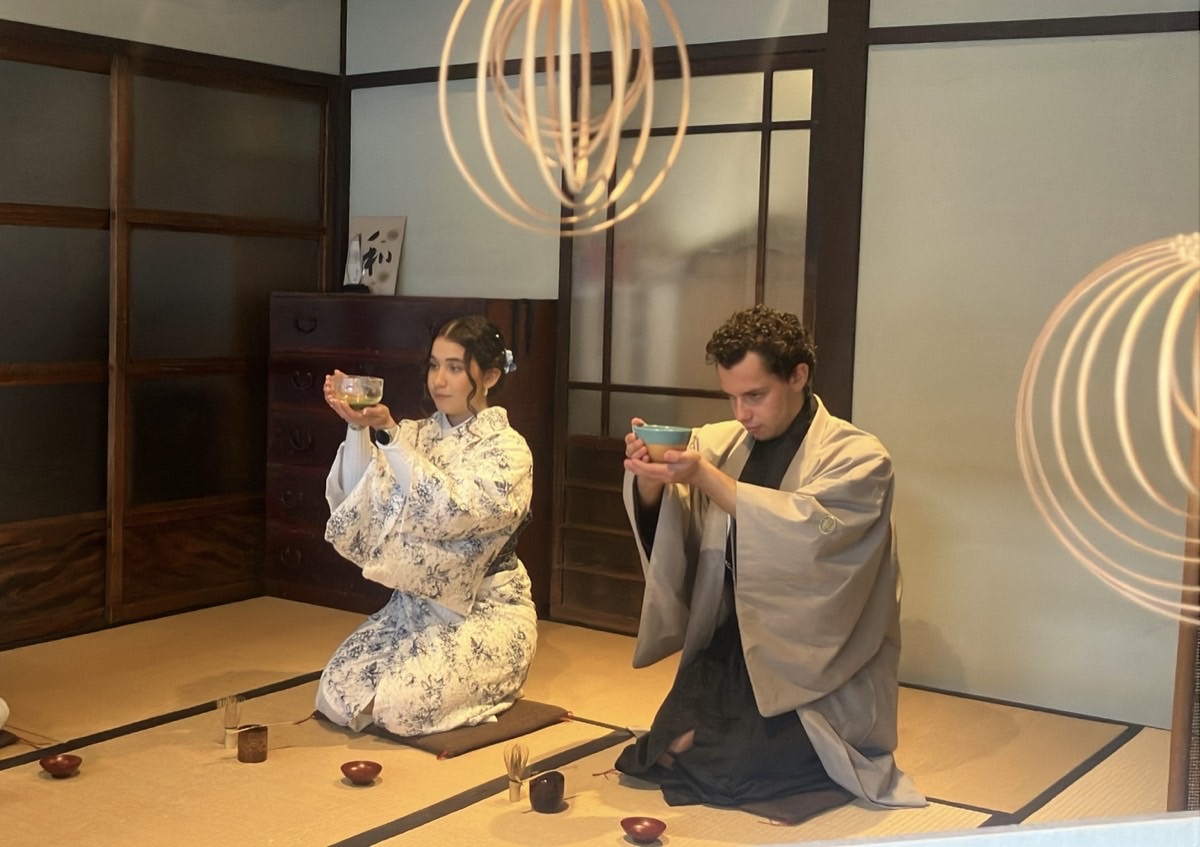 Two guests in kimono kneel and raise glass tea bowls with both hands, beneath woven bamboo spheres hanging from the ceiling.