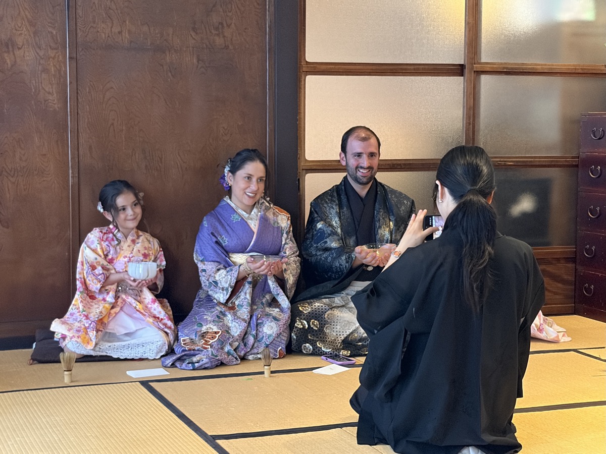 A young guest whisks matcha in a bowl on the tatami while another guest watches beside her.