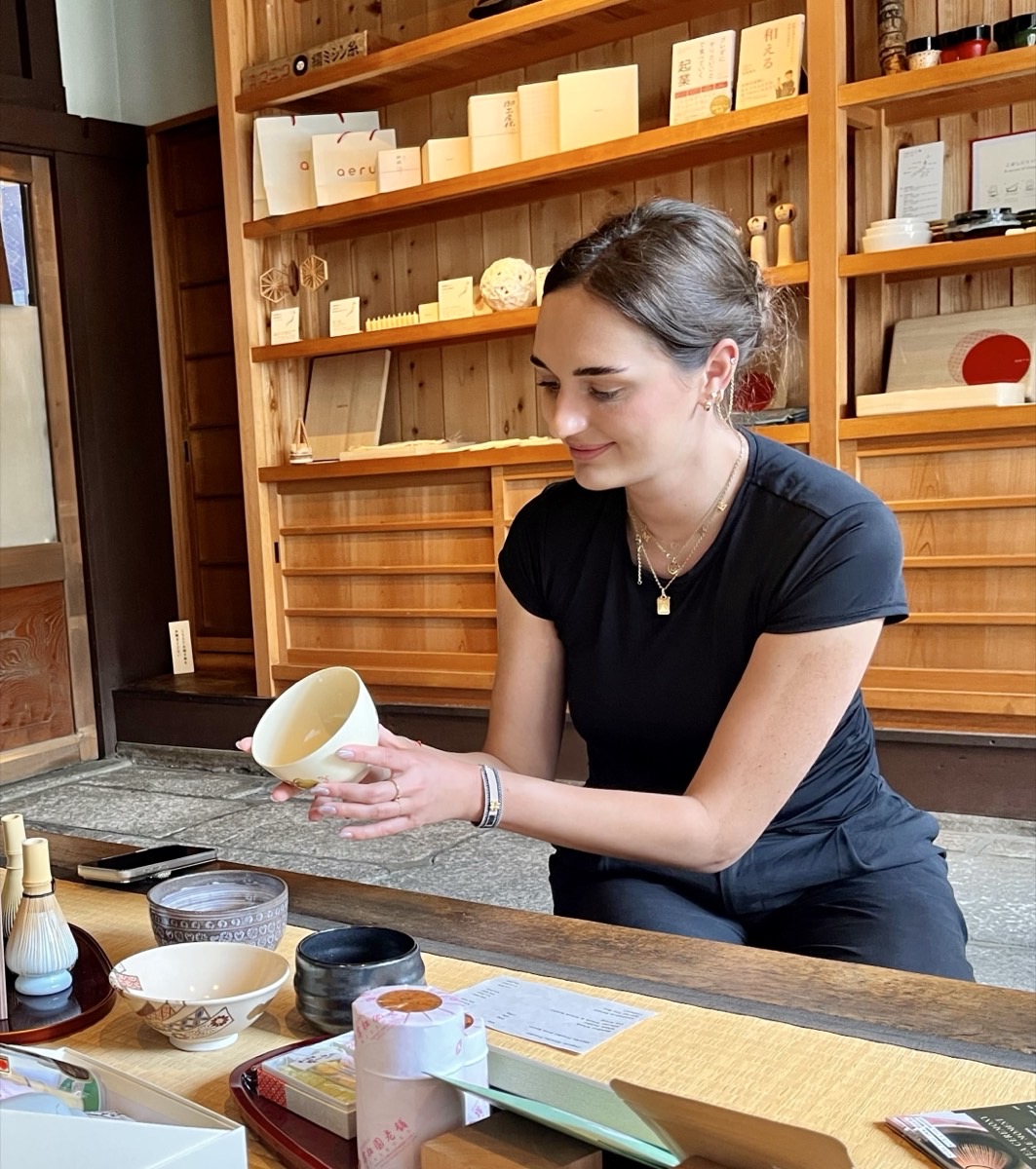 A guest holds a pale tea bowl in both hands, looking at it, in front of shelves of crafts at aeru gojo.