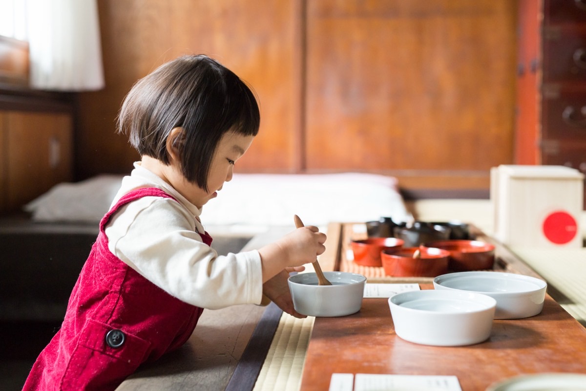 A small child in a red pinafore stirs a bowl with a wooden spoon at a low wooden counter.