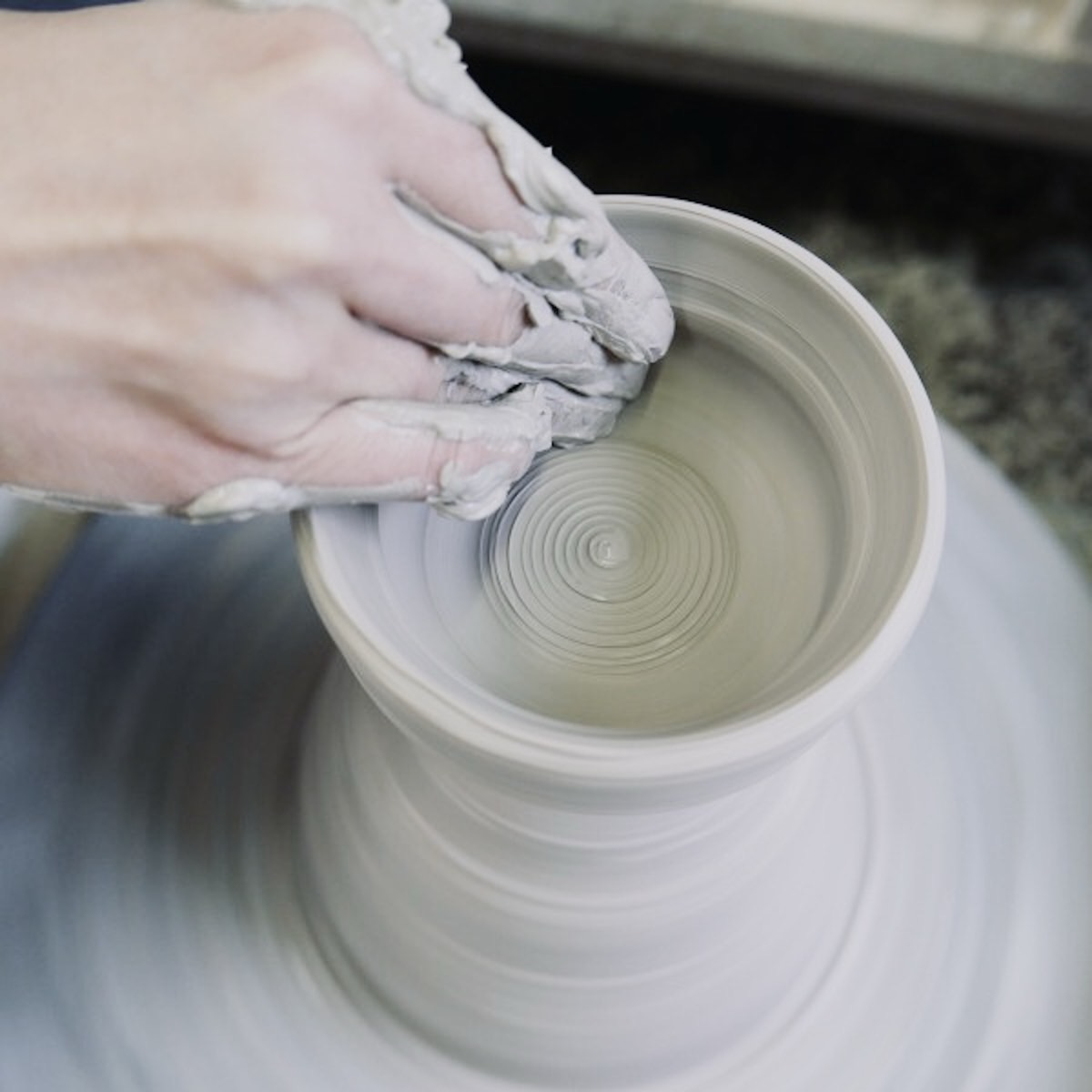 An artisan's clay-covered hands shaping a bowl on a turning potter's wheel.